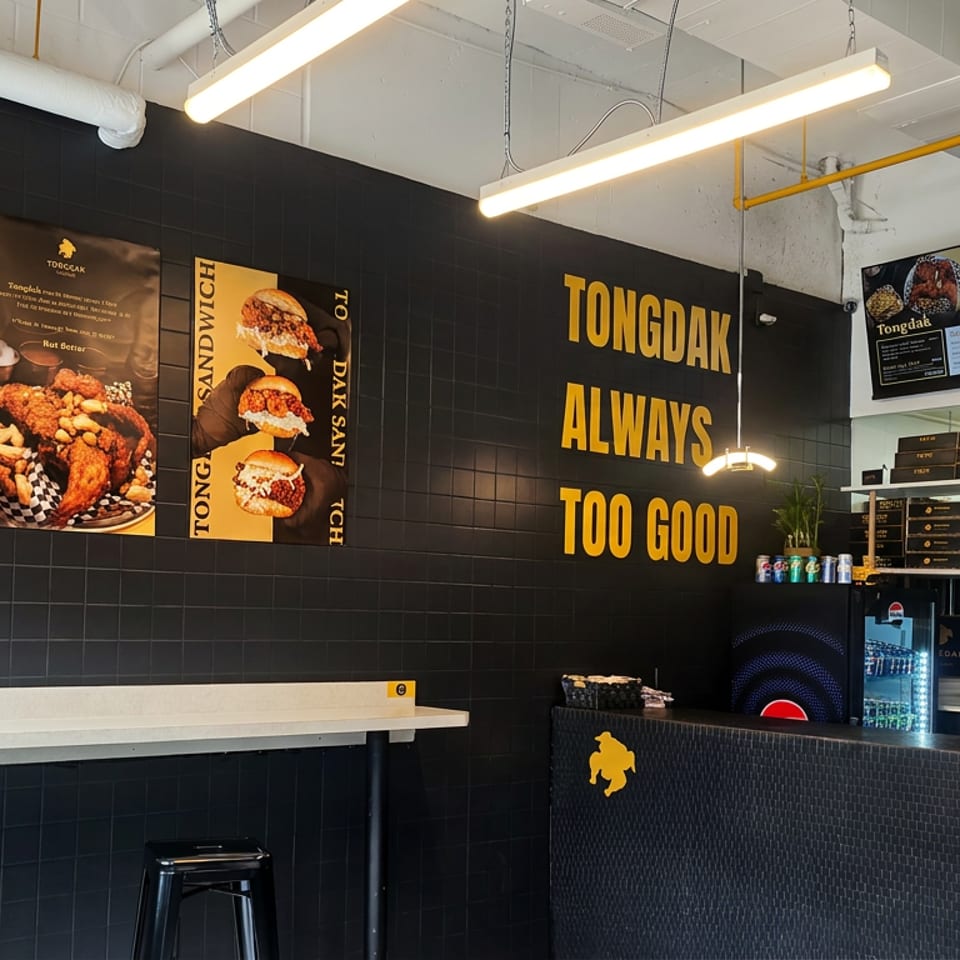 Modern Tongdak Korean fried chicken restaurant interior with black tiled walls, food posters, illuminated menu screens, and ordering counter.