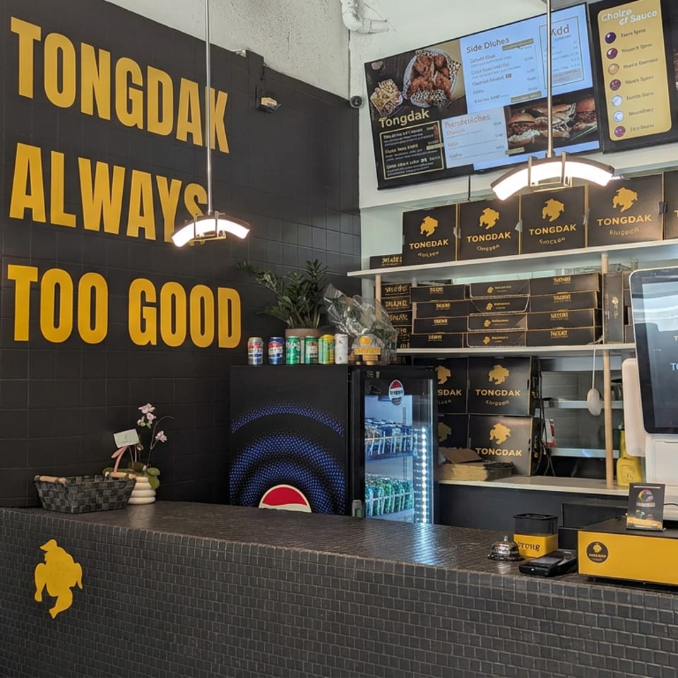 Tongdak restaurant counter with black-and-yellow branding, menu screens, boxed food, beverage refrigerator, and self-ordering kiosk.