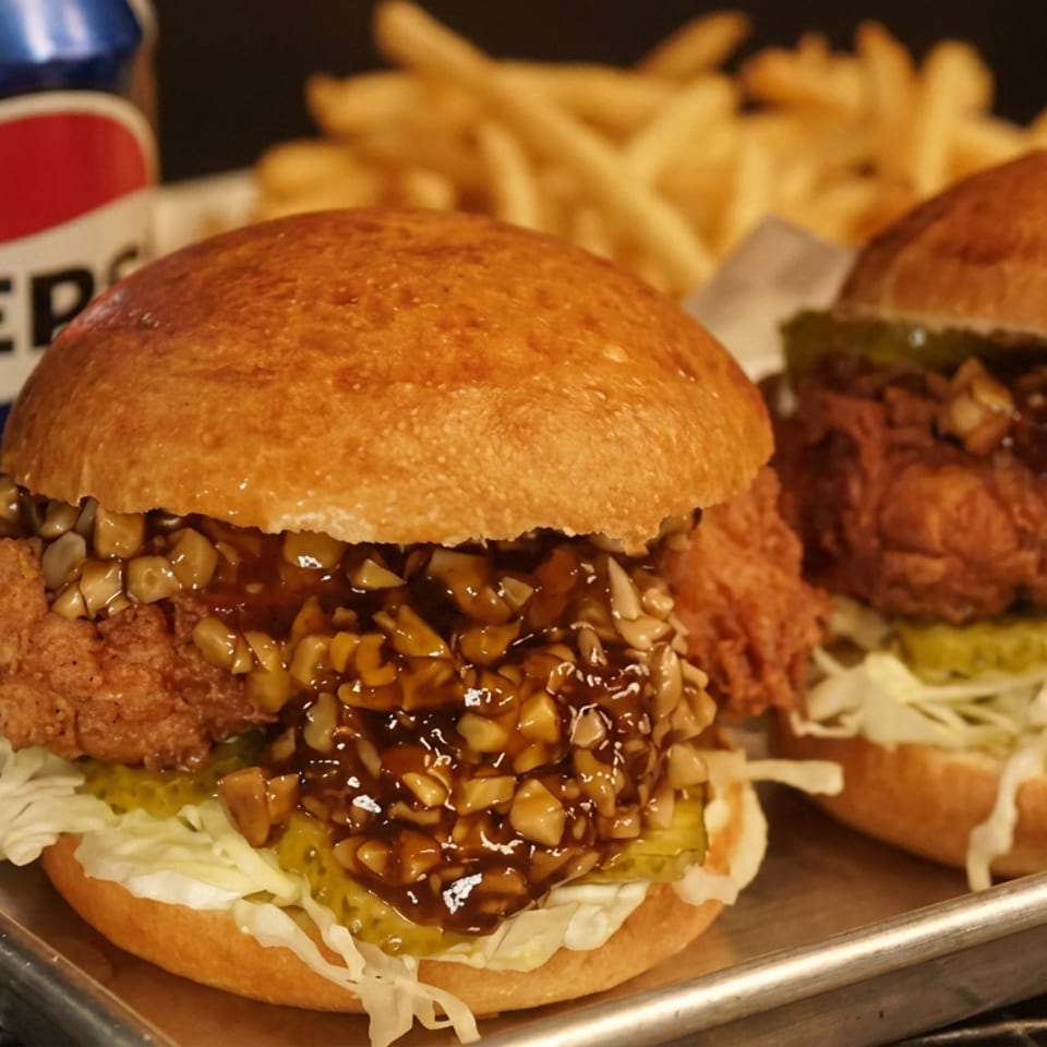 Two crispy chicken burgers topped with glossy sauce and chopped nuts, served with fries and Pepsi on a metal tray.